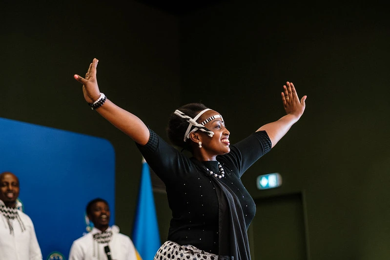 Une danseuse en tenue traditionnelle rwandaise, bras grands ouverts, sur scène devant le drapeau du Rwanda, lors d'Umuganura 2026